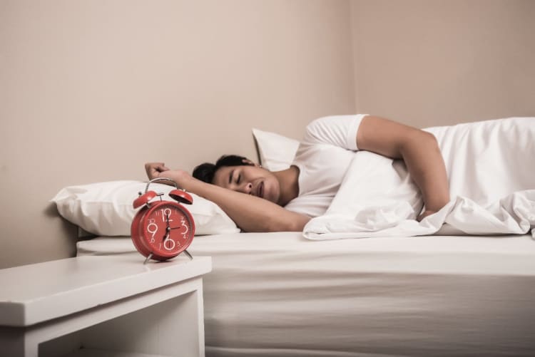 Schlafbedarf-Rechner: Optimale Zu-Bett-Zeit nach Schlafzyklen Mann schläft im Bett, daneben ein Wecker auf dem Nachttisch